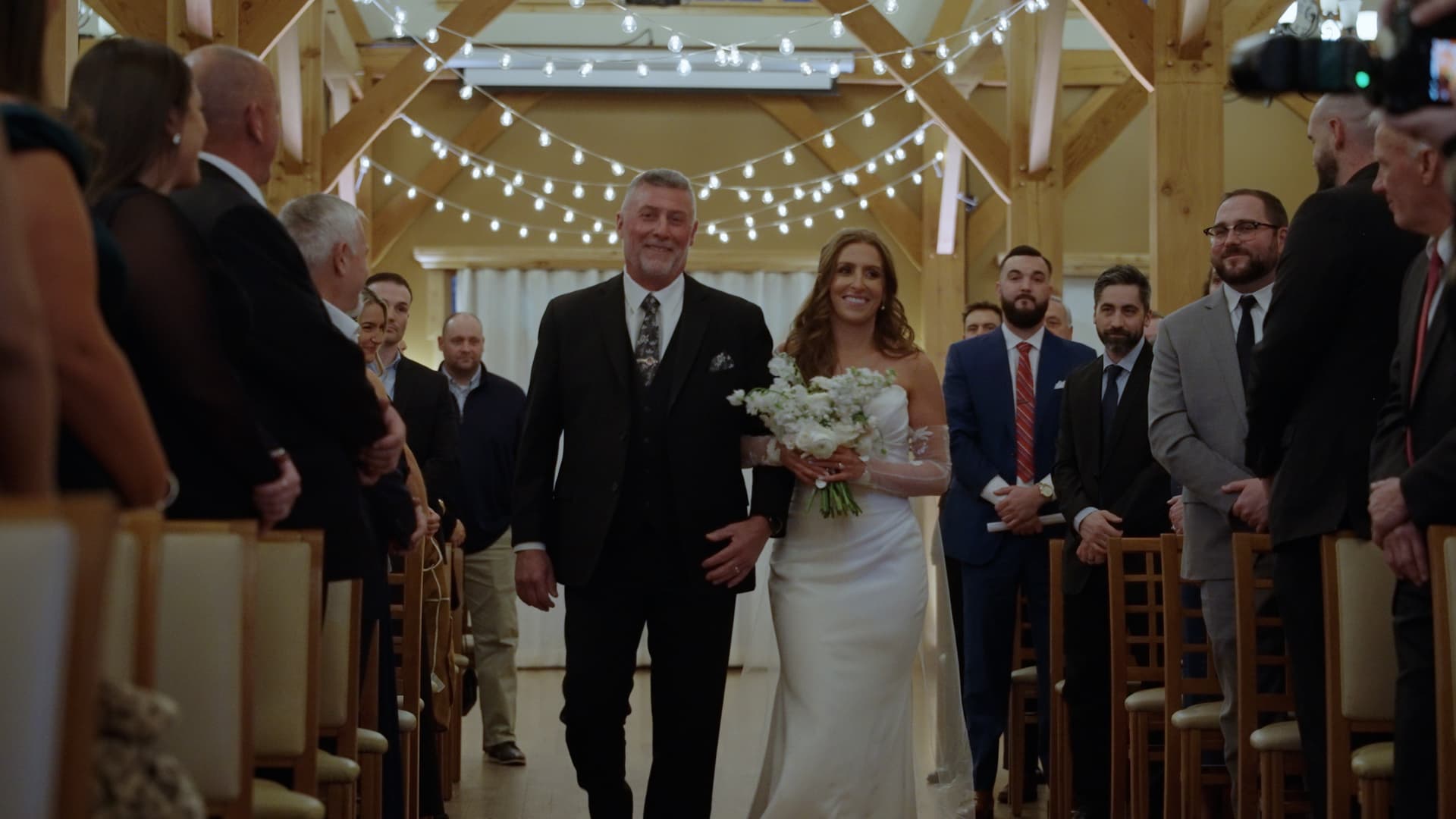A bride walking down the aisle in a string-lit New England barn, by Skytona